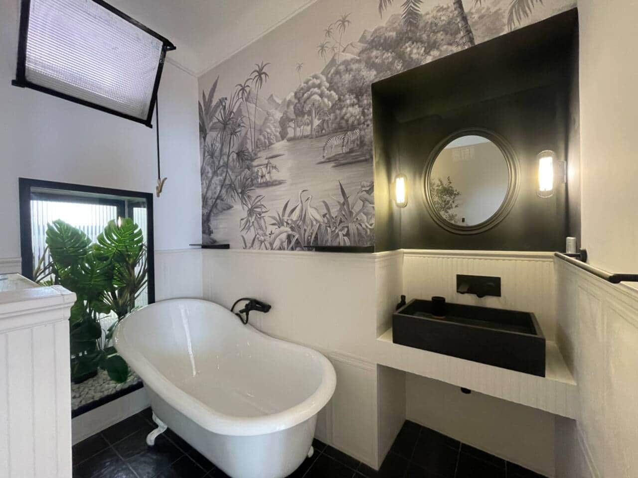 Bathroom with a white clawfoot bathtub against botanical wallpaper of palms and herons, a black stone basin under a round mirror, and a planted alcove behind glass