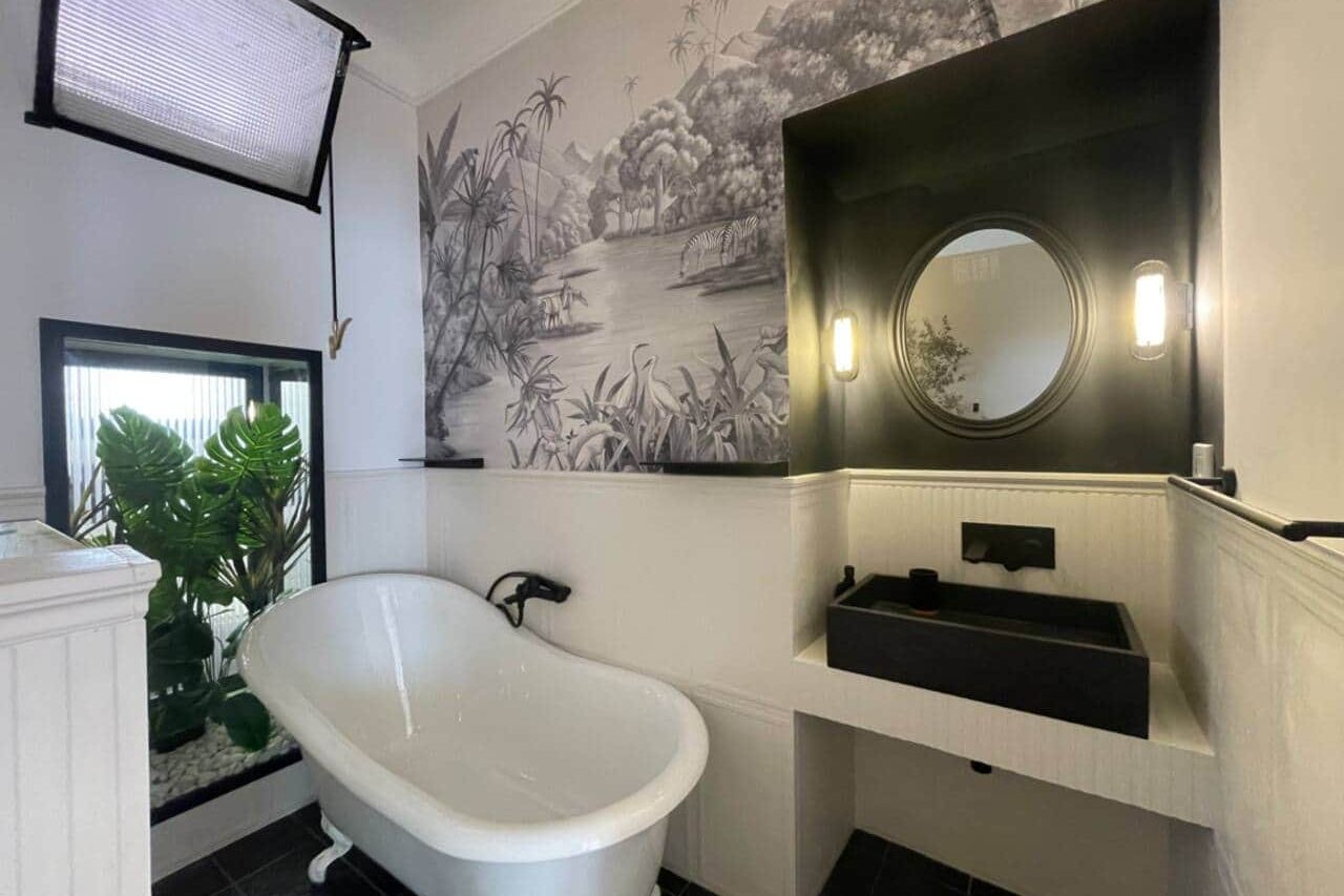 Bathroom with a white clawfoot bathtub against botanical wallpaper of palms and herons, a black stone basin under a round mirror, and a planted alcove behind glass