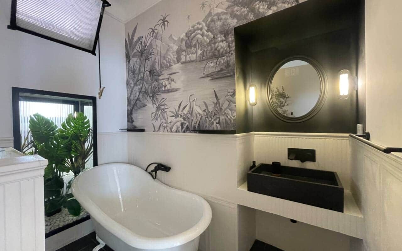 Bathroom with a white clawfoot bathtub against botanical wallpaper of palms and herons, a black stone basin under a round mirror, and a planted alcove behind glass