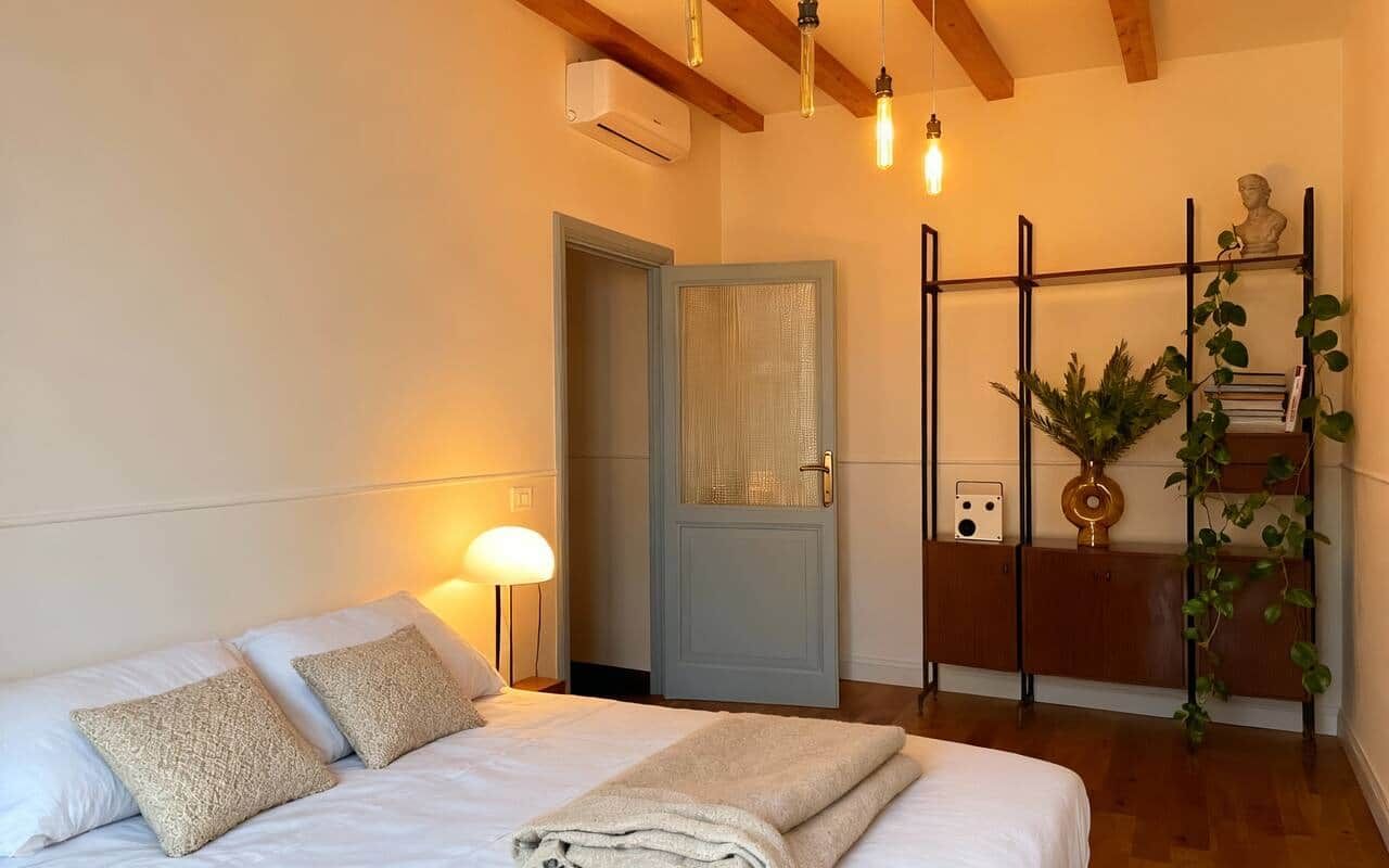 The same bedroom by lamplight, with a wall-mounted air conditioning unit, a glazed grey door, and black shelving holding plants, books and a small bust