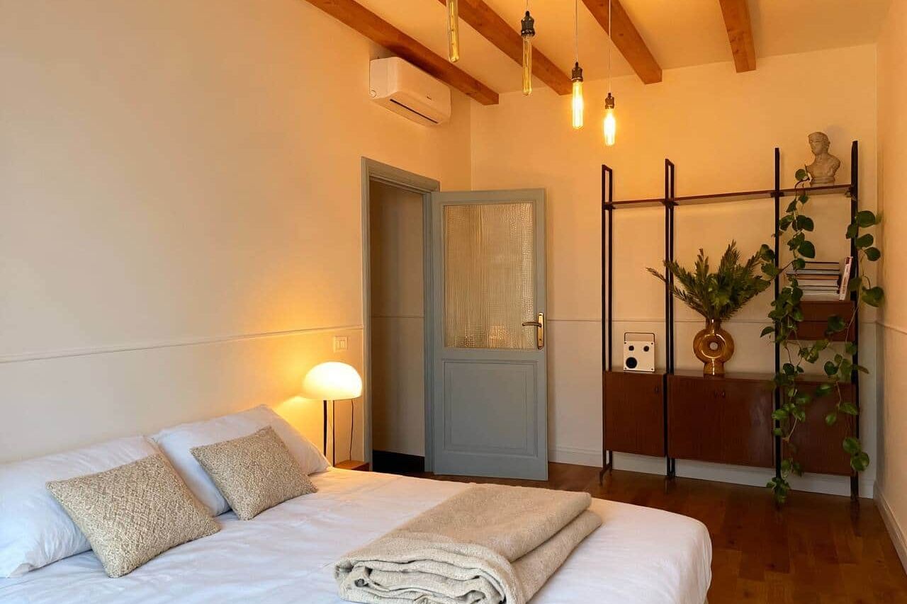 The same bedroom by lamplight, with a wall-mounted air conditioning unit, a glazed grey door, and black shelving holding plants, books and a small bust