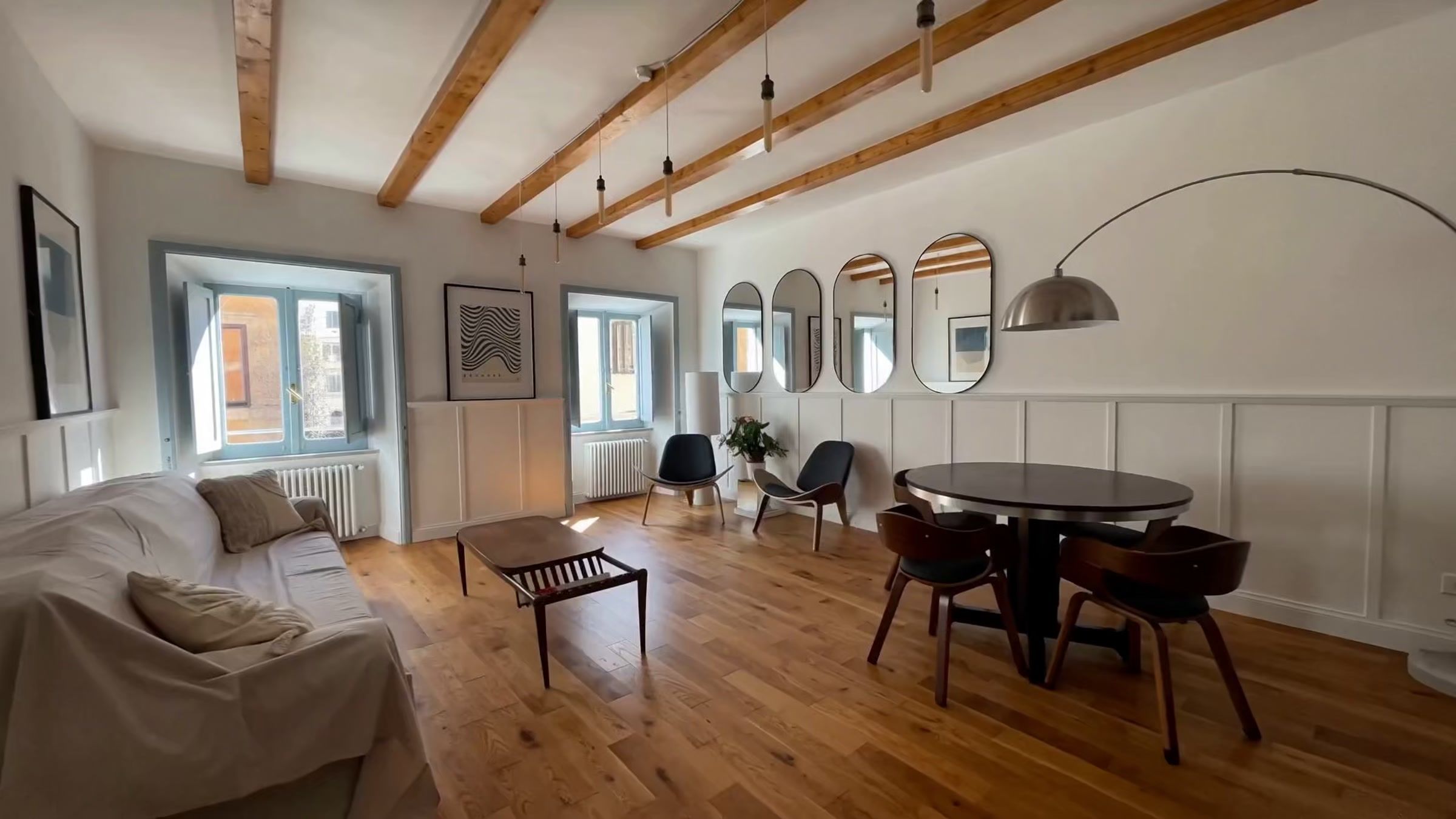 The living room seen from the door: exposed timber beams, a long dining table, an arc lamp and a row of oval mirrors along the wall