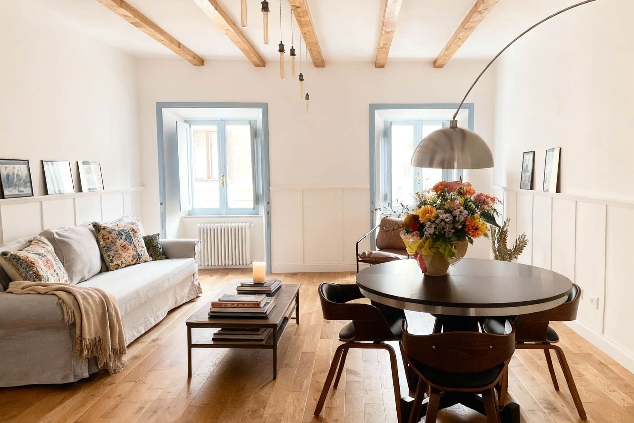 Living room with a pale linen sofa, stacked books on a low table, a round dining table with a vase of flowers, and two shuttered windows in blue-grey frames