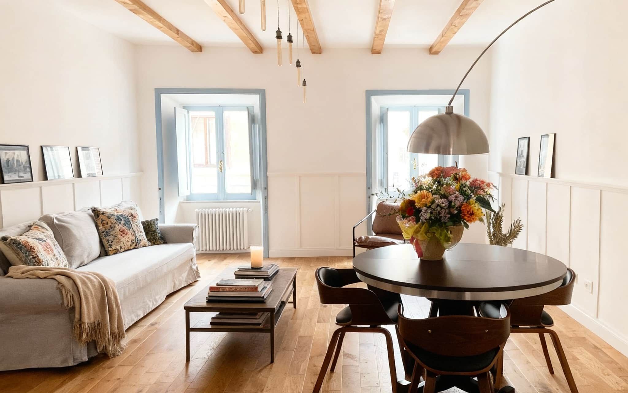 Living room with a pale linen sofa, stacked books on a low table, a round dining table with a vase of flowers, and two shuttered windows in blue-grey frames